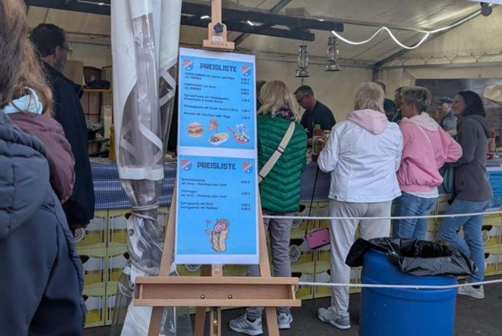 Menschenschlange an einer Theke zur Essensausgabe, an der die Menüs und Preise angeschlagen sind.