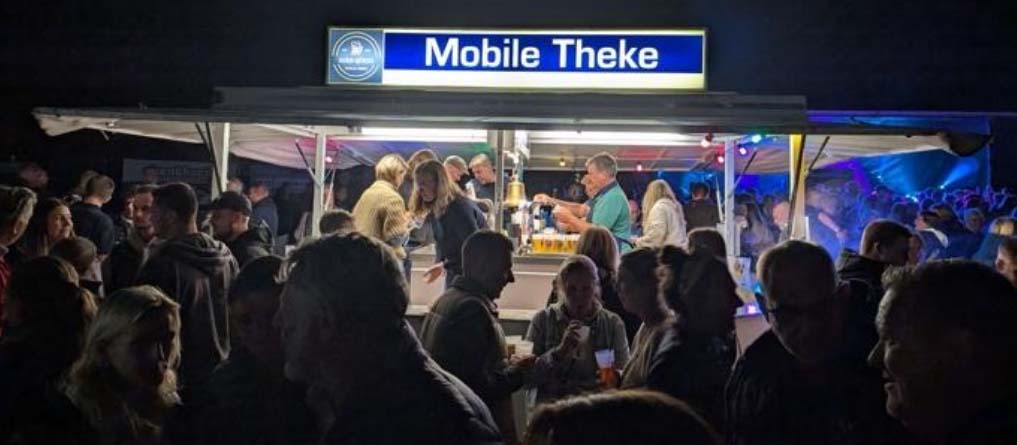 Von Menschen umringter Getränkestand mit der Leuchtschrift “Mobile Theke” in der Dunkelheit.