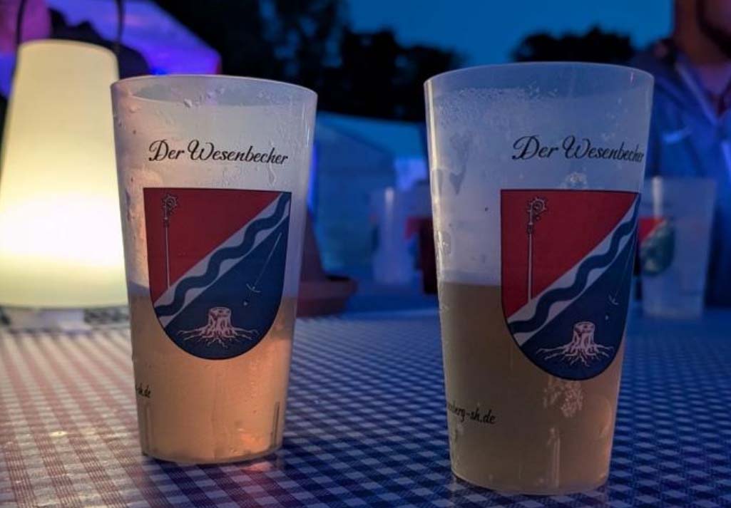 Getränkebecher mit Aufschrift „Der Wesenbecher” und Logo der Gemeinde auf einem Stehtisch.