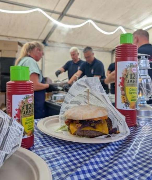 In Papiertüte eingepackter Burger auf dem Tresen. Im Hintergrund die Küchenhelferinnen und -helfer.