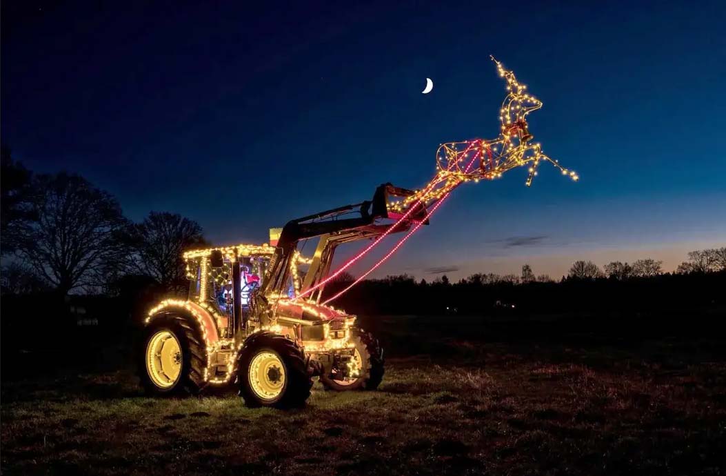 Auf einer Wiese im Mondschein steht ein mit Lichterketten erleuchteter Traktor, der an seiner erhobenen Schaufel ein aus Lichtern gebildetes Rentier trägt.